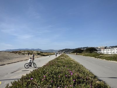 Wide road flanked by vegetation and sand dunes under a clear blue sky. A child rides a bicycle on a paved path. Hills and buildings are visible in the background.