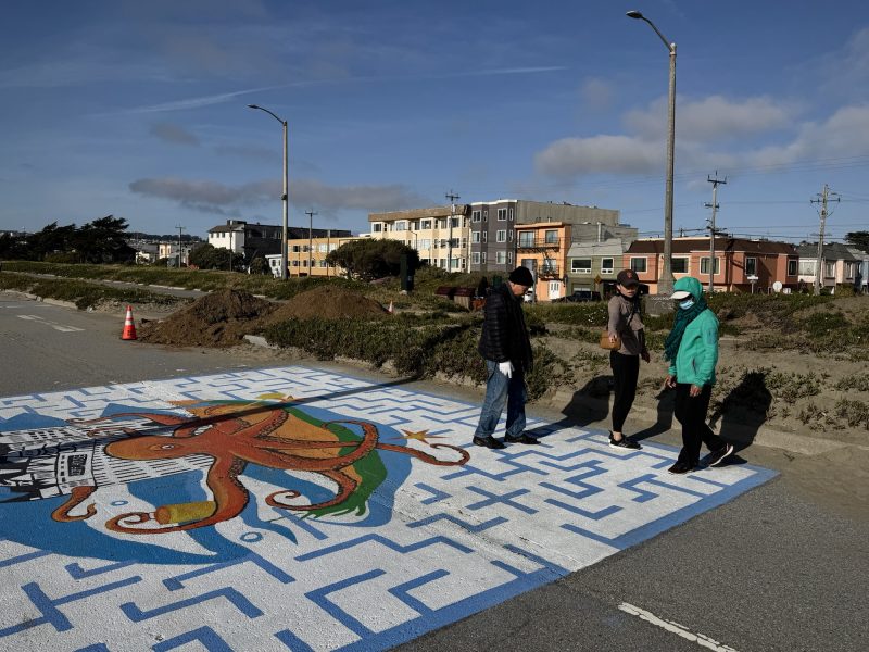 Three people in jackets and hats stand on a painted street mural featuring an octopus and skyline, with houses and a construction cone in the background.