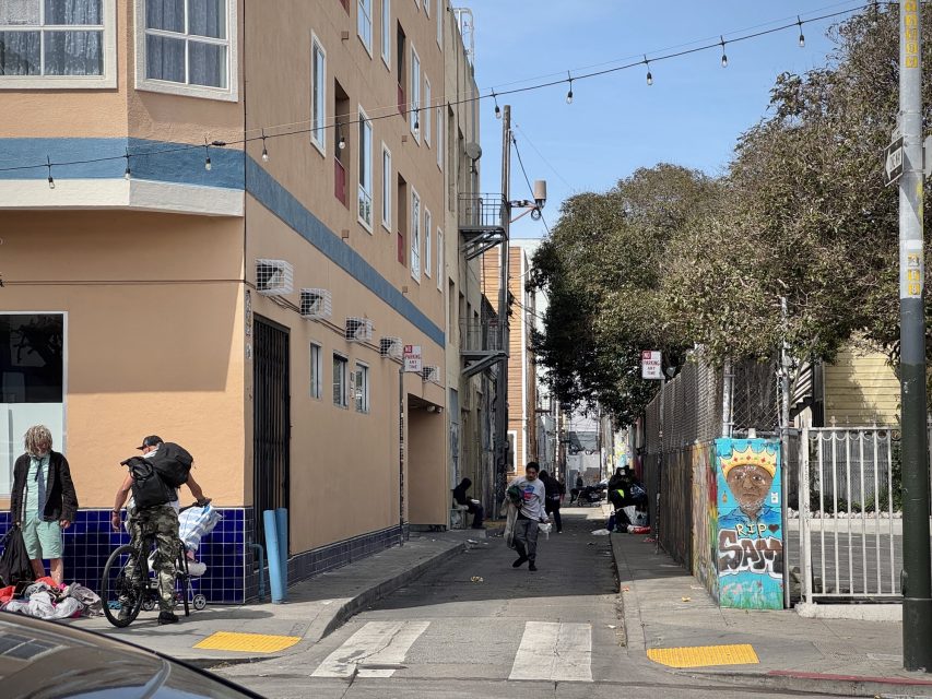 Urban alleyway with people, bicycles, and graffiti. Trees and a peach-colored building line the street. Pedestrians walk and gather near a fence. String lights hang above.