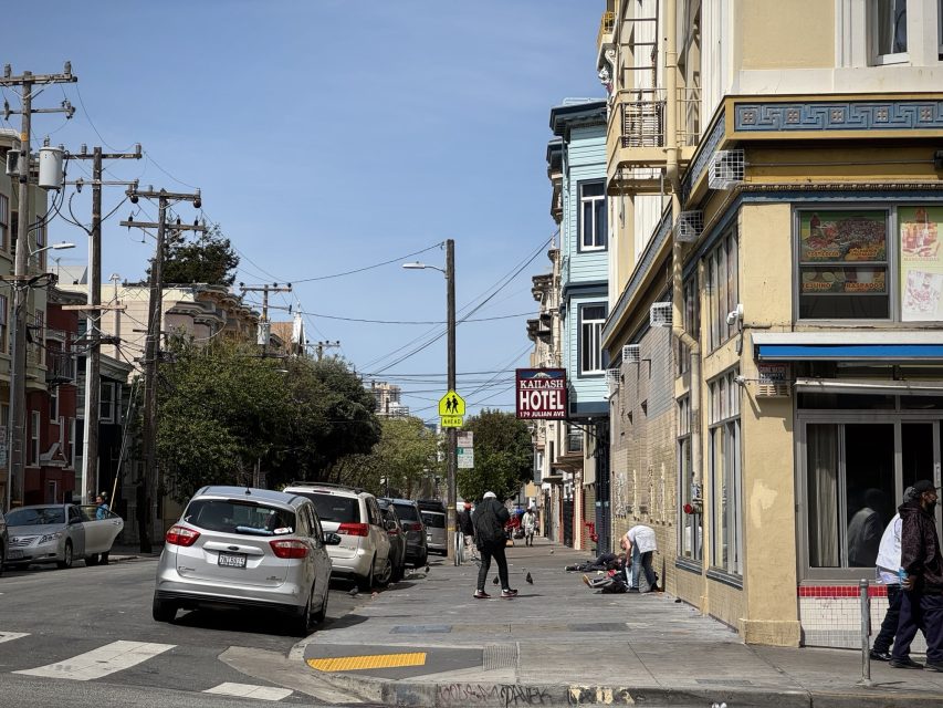 City street scene with parked cars, pedestrians, and buildings. A hotel sign is visible in the distance. People are walking and interacting on the sidewalk.