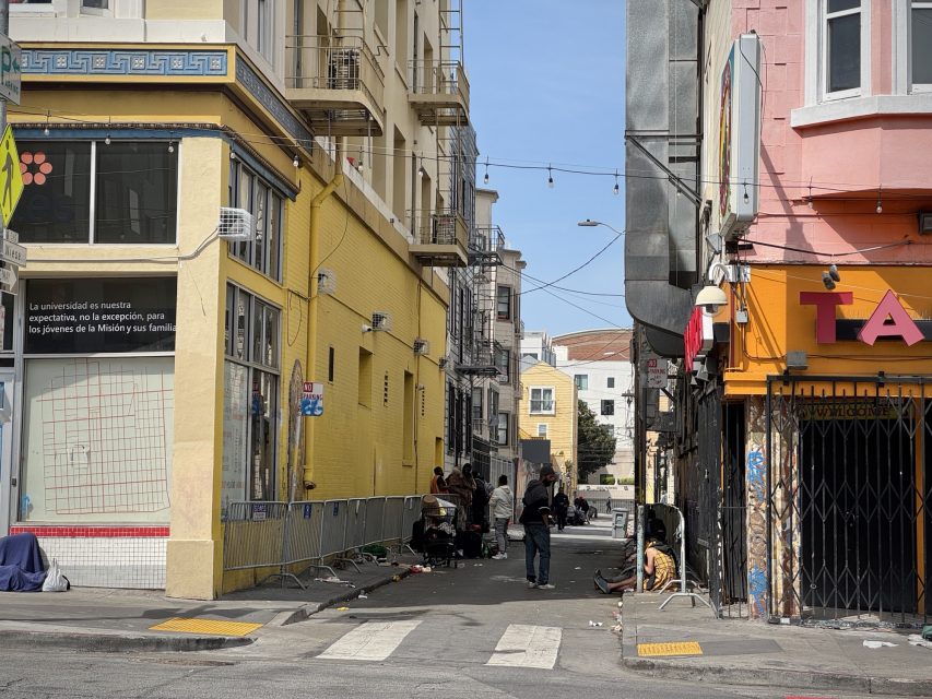 Urban alleyway with several people gathered, surrounded by colorful buildings. A mural with Spanish text is on the left wall. Some string lights hang above. Street signs and debris visible.