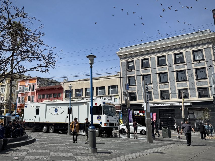 Truck parked in a city square beside a row of colorful buildings with a few people walking nearby. Pigeons fly overhead.