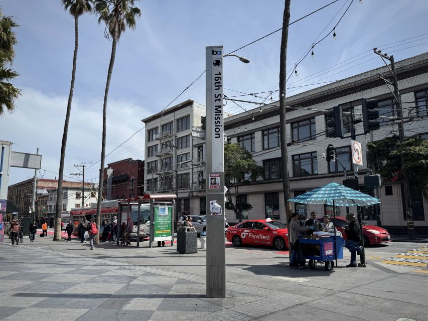 Urban street scene at 16th St Mission intersection, featuring buildings, people walking, a trolley stop, red vehicles, and food stalls under umbrellas in a cityscape.