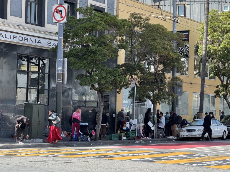 A group of people gathered on a city sidewalk near a building with "California" on it. Some appear to be seated, with belongings nearby. Traffic signs and trees are visible.