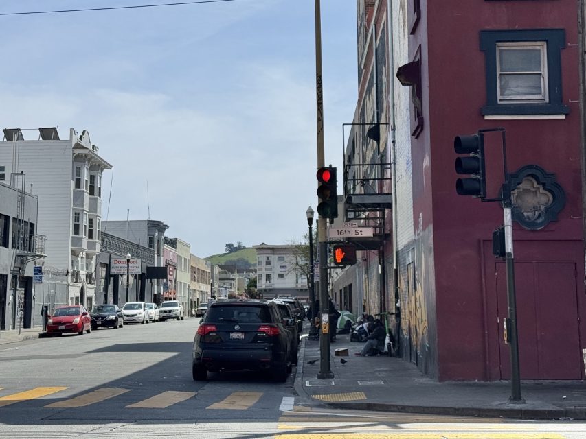 Street scene with cars parked along the sidewalk, a red traffic light, and pedestrians sitting near a red building. Urban environment with buildings extending into the background.