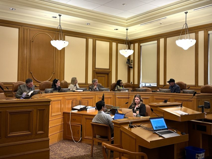 A group of people seated in a formal meeting room with wood paneling, using laptops and papers.