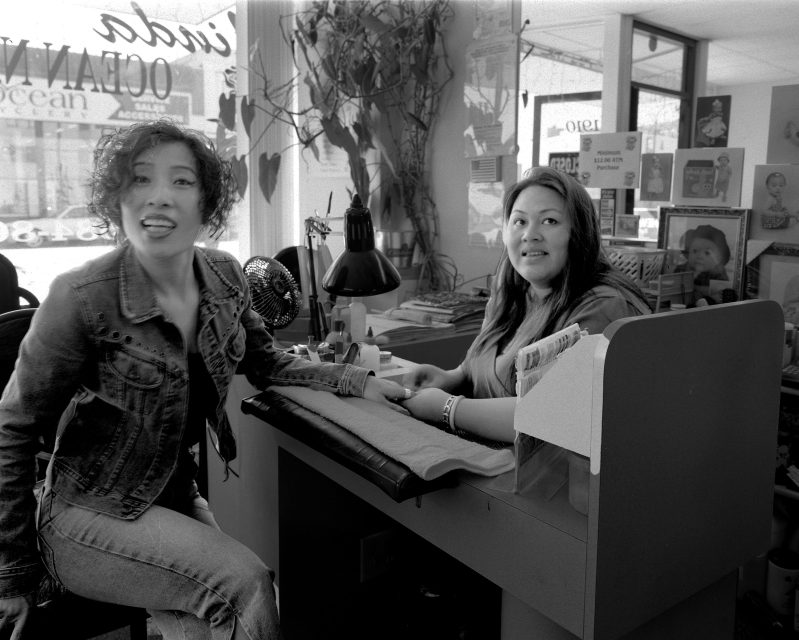 Two women in a nail salon; one woman sits at a manicure table smiling, the other sits across, playfully sticking out her tongue, with salon supplies and décor visible in the background.