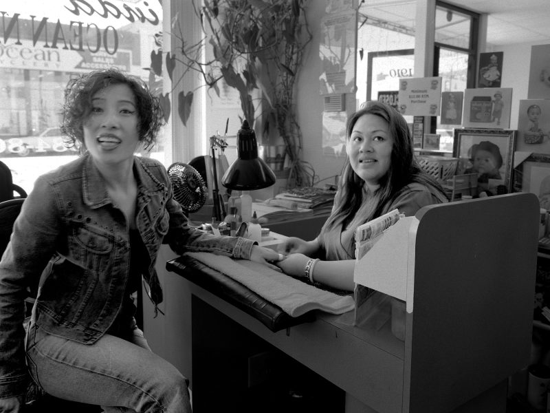 Two women in a nail salon; one woman sits at a manicure table smiling, the other sits across, playfully sticking out her tongue, with salon supplies and décor visible in the background.