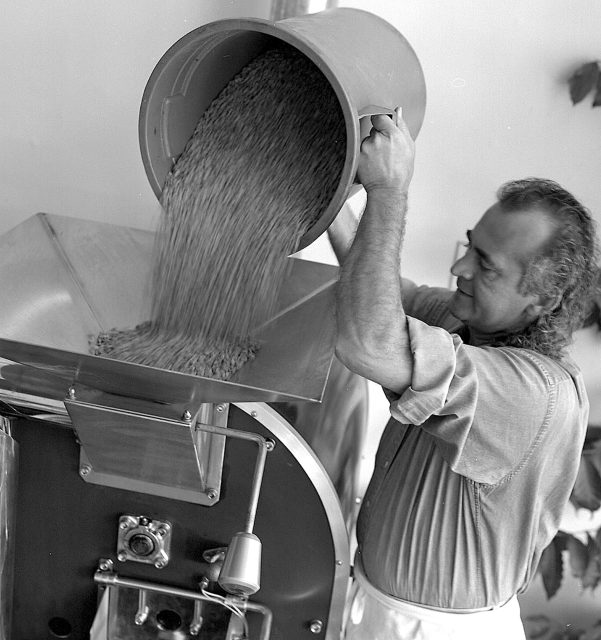 A man pours a large container of coffee beans into a commercial coffee roasting machine.