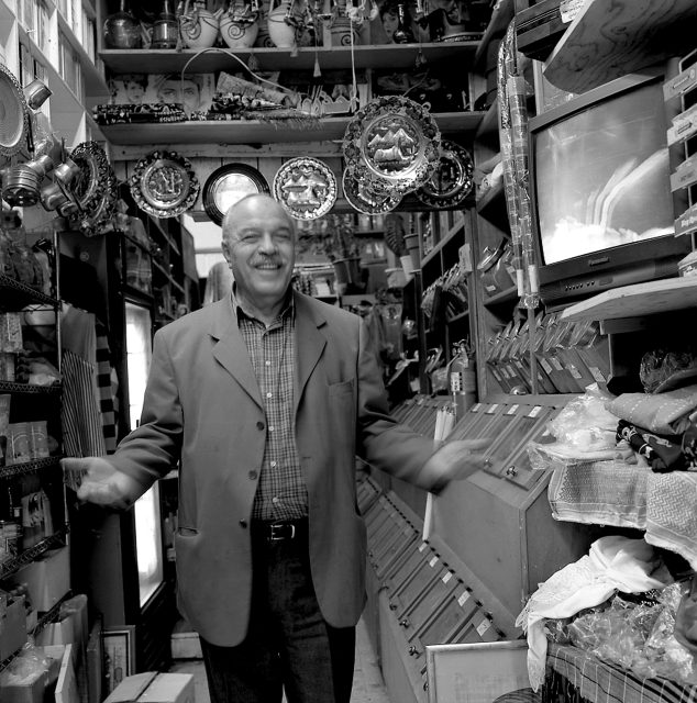 A man in a suit jacket stands smiling inside a small, crowded shop filled with plates, trinkets, and a television.