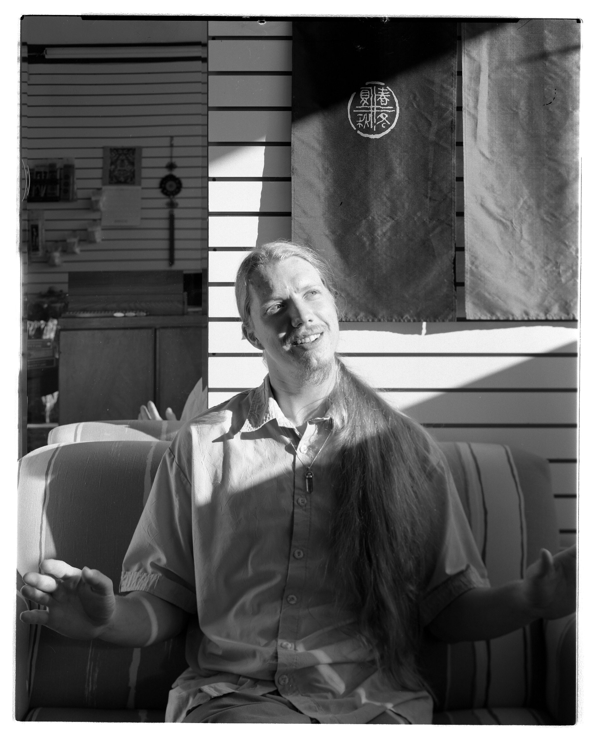 A person with long hair sits on a striped couch, gesturing with both hands. Sunlight and shadows fall across their face and body. A dark cloth with Asian characters hangs behind them.