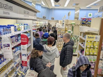 Shoppers wait in a long line inside a pharmacy, standing between aisles of health products and personal care items.