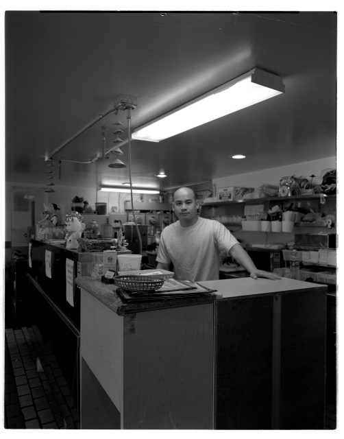 A person stands behind the counter in a small kitchen or food service area, surrounded by shelves with various containers and supplies.