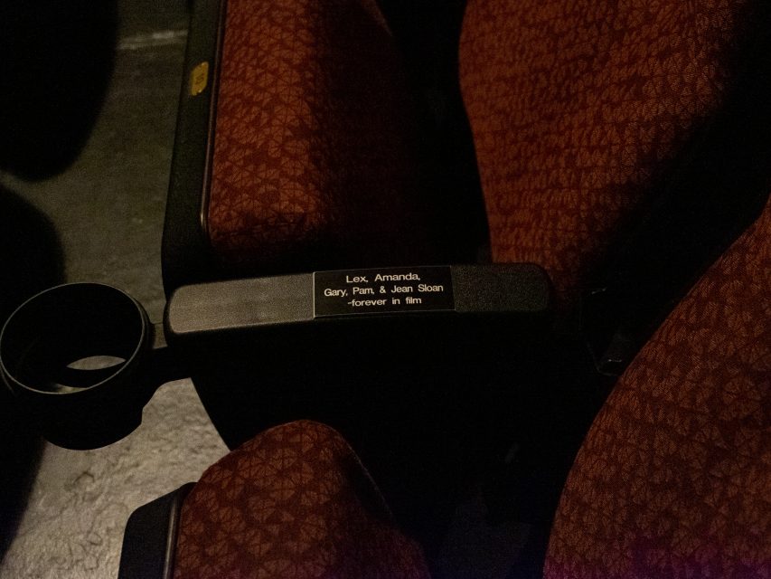 Close-up of a red-patterned theater seat with a black armrest that has a plaque reading, "Lex, Amanda, Gary, Pat, & Jean Sloan - Forever in film.
