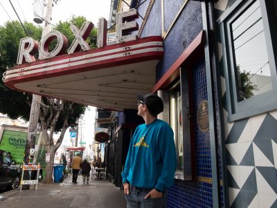 A person in a blue sweatshirt stands in front of the Roxie theater marquee, looking up at the sign on a city sidewalk.