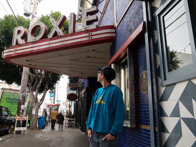 A person in a blue sweatshirt stands in front of the Roxie theater marquee, looking up at the sign on a city sidewalk.