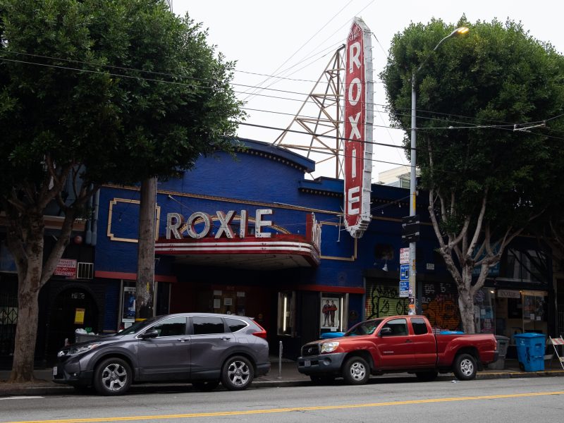 Street view of the Roxie Theater, a blue building with a large vertical and marquee sign, flanked by parked cars and trees.