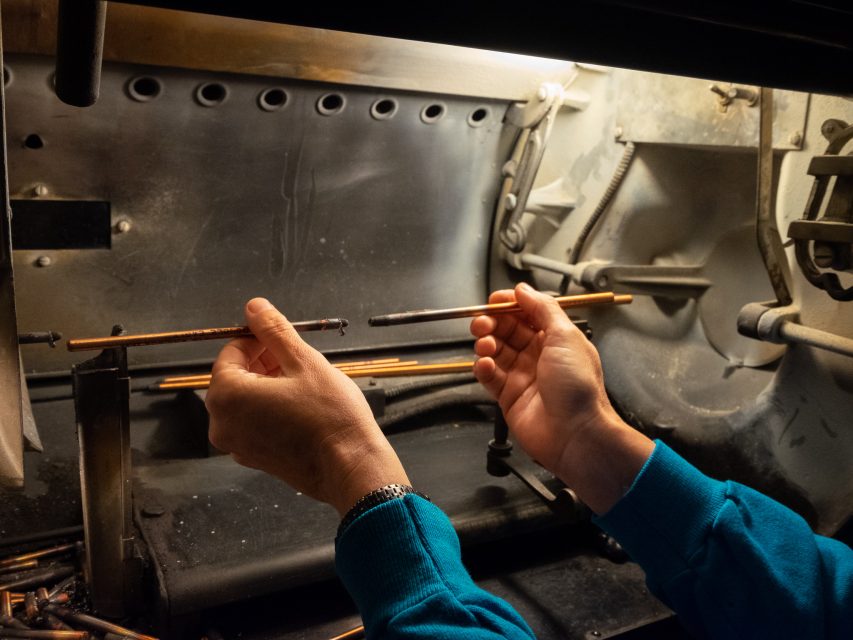 A person holds two rods inside a machine, comparing a clean rod to a dark, sooty one in an industrial or workshop setting.