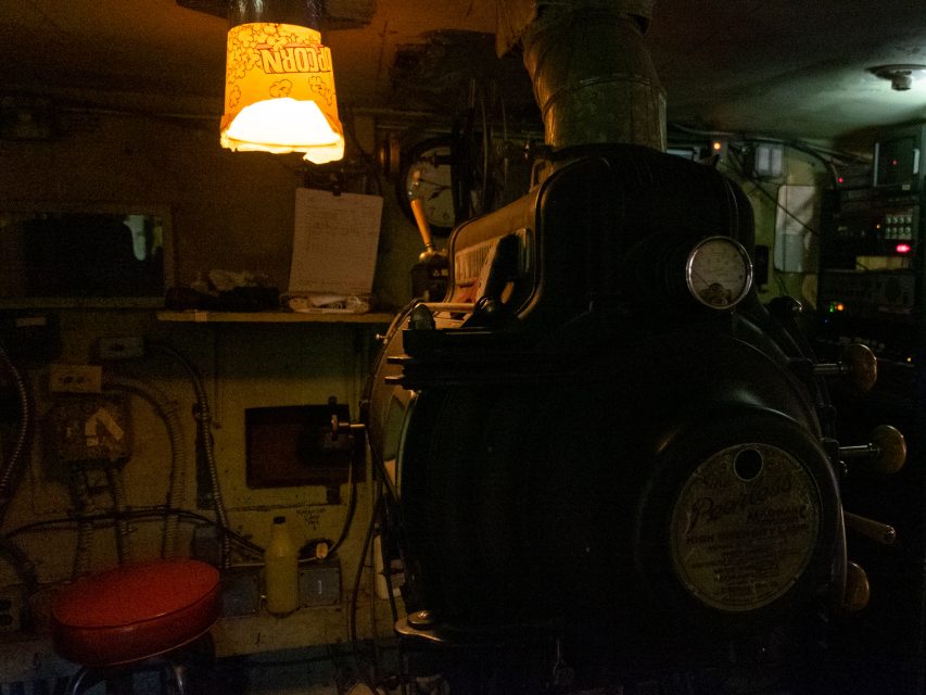 A dimly lit room with industrial equipment, a red stool, control panels, exposed wiring, and a single overhead lamp with a yellow shade.