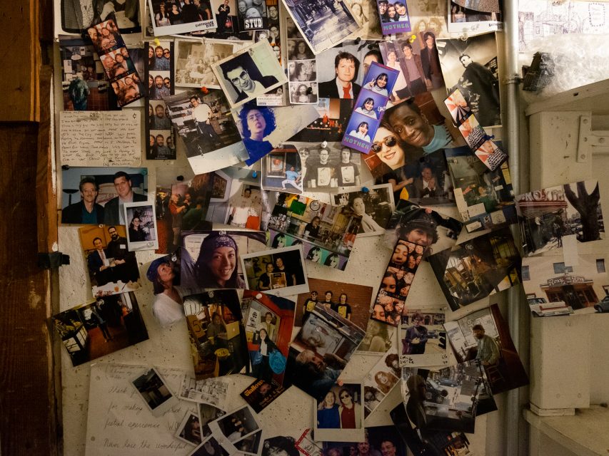 A cluttered bulletin board covered with numerous candid and posed photographs of people, Polaroids, handwritten notes, and small mementos, creating a collage-like display.