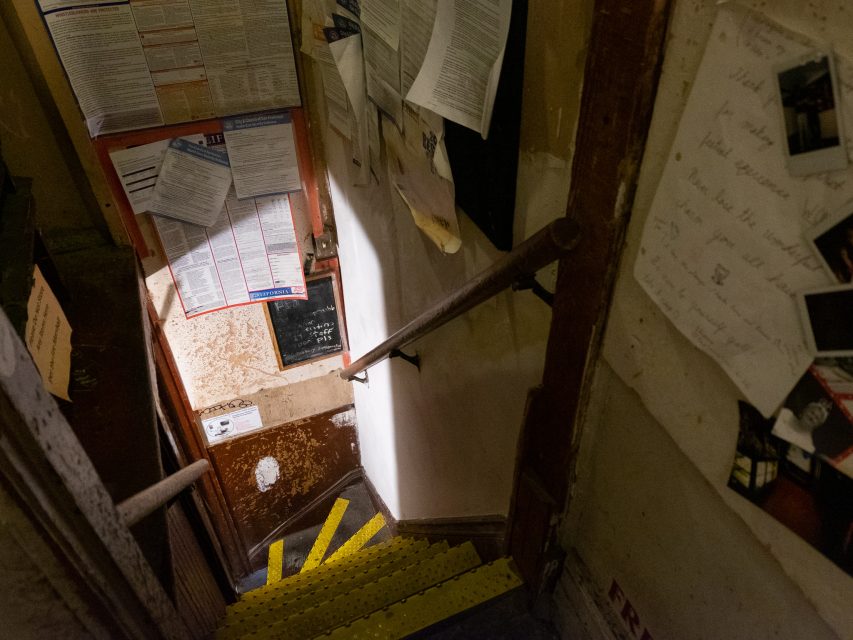 A narrow, dimly lit stairwell with yellow-striped steps and various papers and notices posted on the surrounding walls.