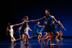 A group of dancers in blue and white outfits perform barefoot on stage, holding hands in a coordinated formation against a dark blue background.