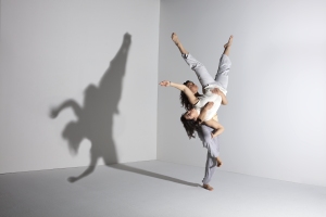 Two dancers in light attire perform a dynamic lift in a minimalist setting, casting a dramatic shadow on the wall.