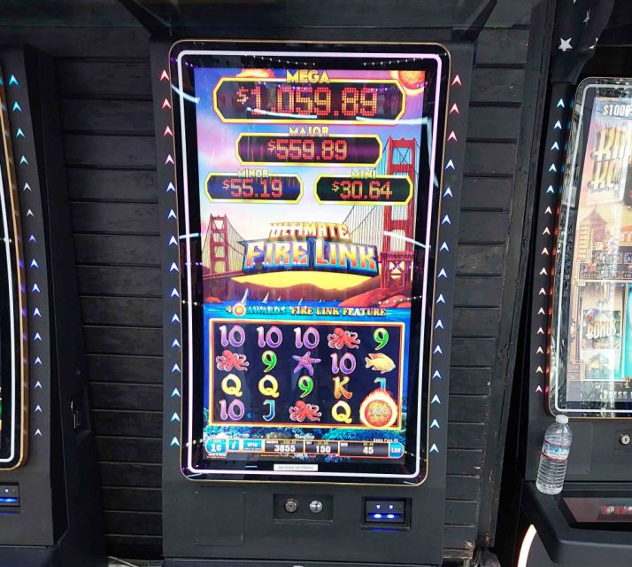 Slot machine with "Ultimate Fire Link" game displayed. Bright colors, jackpot amounts shown at top. Numbered and themed symbols are visible. Lit buttons and water bottle nearby.