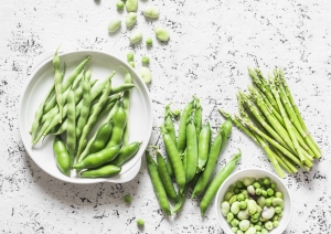 Variety of green vegetables on a white surface: a plate of snap peas, loose broad beans, a bunch of asparagus, green peas and beans in a bowl.
