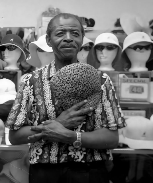 A man stands in front of a hat display, holding a patterned hat to his chest. Mannequin heads with hats and sunglasses fill the background.