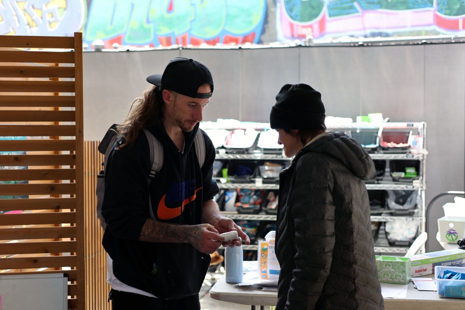 A man in a black cap and backpack hands an item to a woman in a coat and beanie at an outdoor booth with shelves and supplies in the background.