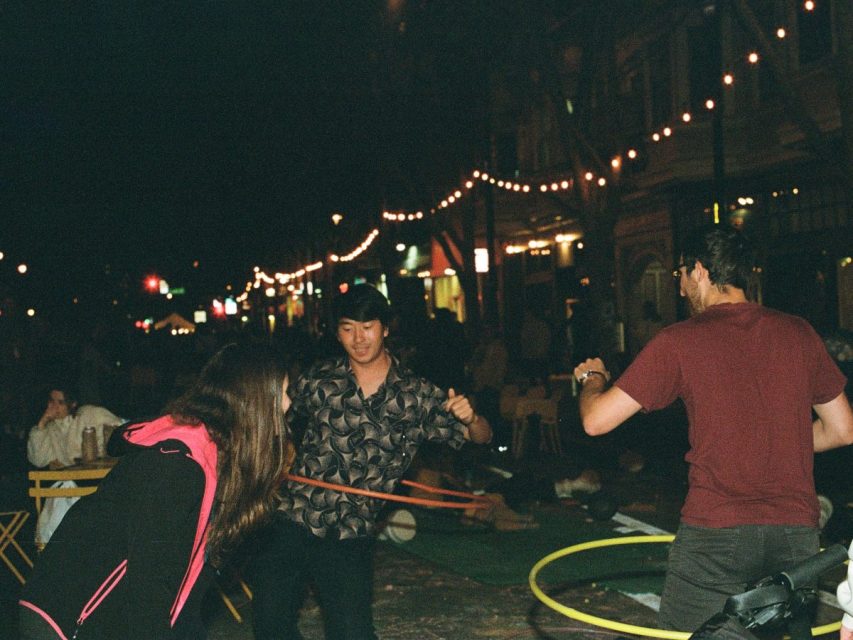 Three people are hula hooping outdoors at night under string lights, with other people seated at tables in the background.