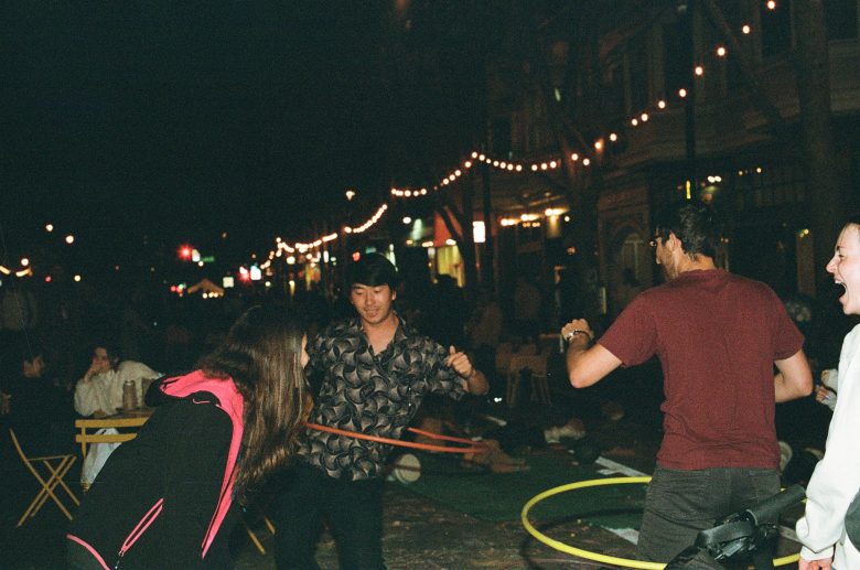 Three people hula hoop at night on a lively, string-lit street, while others sit and watch in the background.
