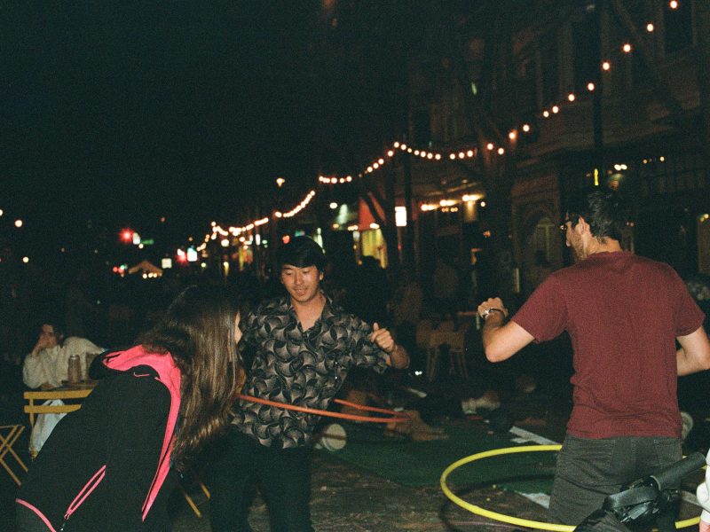 Three people hula hoop at night on a lively, string-lit street, while others sit and watch in the background.