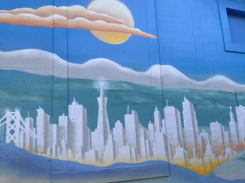 A mural depicts a white city skyline with a tall central tower, distant bridge, hills, clouds, and a yellow sun against a blue background.
