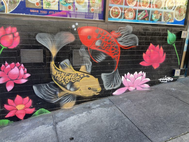 A mural of two koi fish, one red and one yellow, swimming among pink lotus flowers painted on a black brick wall below restaurant menu photos.