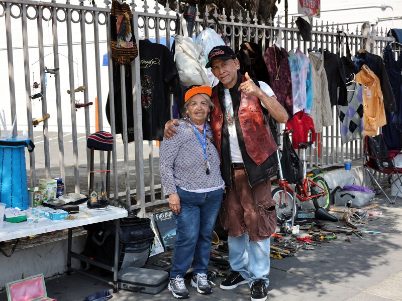 Two people stand smiling together on a sidewalk, surrounded by various clothing items and objects displayed for sale along a metal fence.