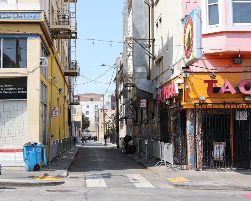 A narrow city alley bordered by yellow and beige buildings, with barricades, trash bins, and a closed taqueria on the right.