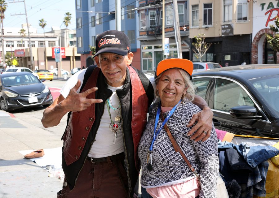 Two people stand on a city sidewalk smiling at the camera; one wears a vest and cap, the other an orange hat, with cars and buildings in the background.