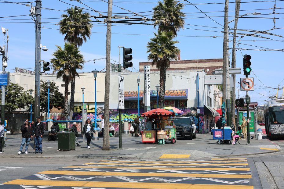 A busy urban street corner with pedestrians, palm trees, a food cart, graffiti-covered buildings, and overhead power lines under a clear sky.