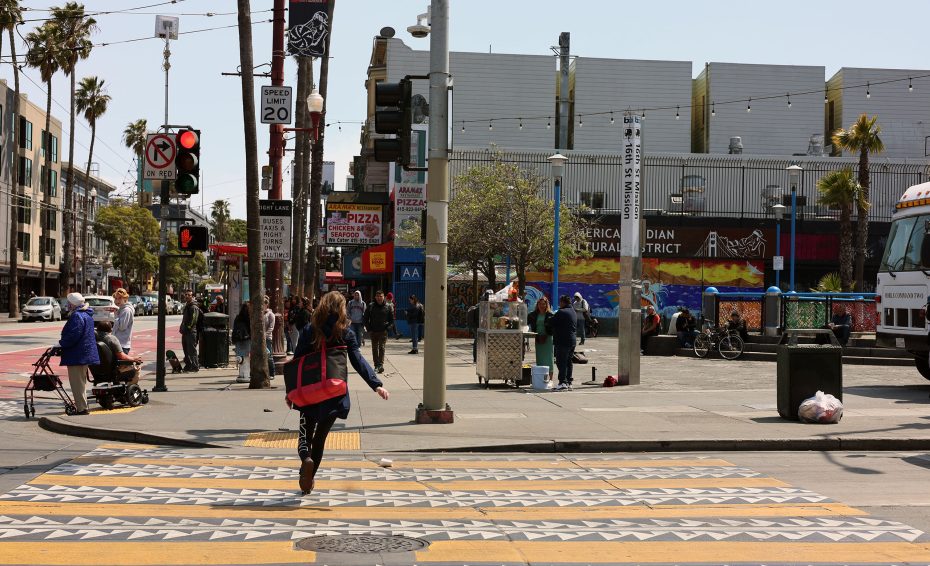 A person with a red bag crosses a city street at a crosswalk; people are gathered on the sidewalk near urban buildings and street art.