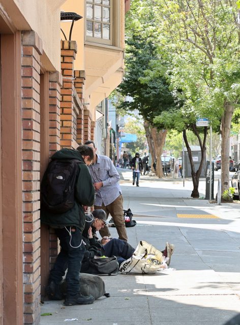 Several people sit and stand on a city sidewalk next to a brick building, surrounded by bags and personal belongings. Trees line the street in the background.