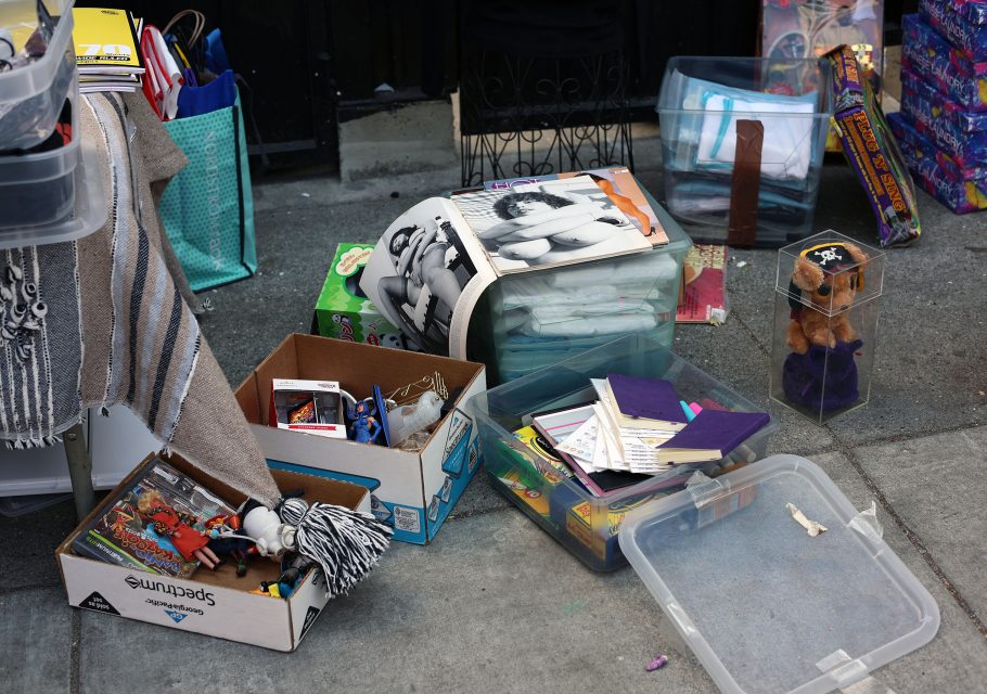 A variety of miscellaneous items in boxes and containers are displayed on a sidewalk, including books, toys, papers, a striped blanket, and a plush bear in a pirate hat.