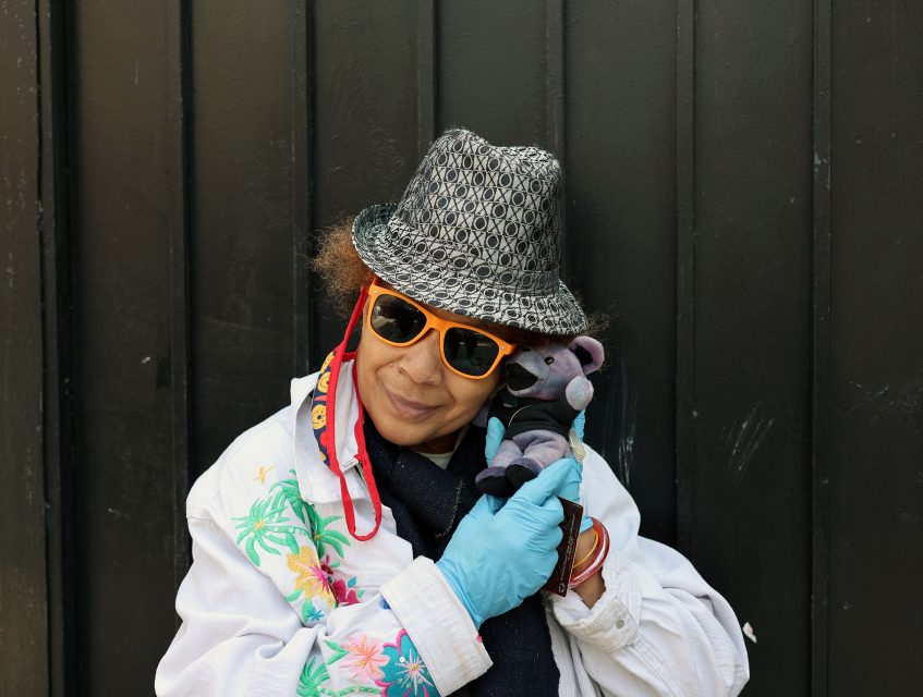 A person wearing a patterned hat, orange sunglasses, blue gloves, and a floral shirt holds a small stuffed bear next to their face, standing in front of a dark background.