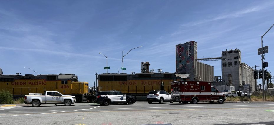 A Union Pacific train passes through an intersection as several vehicles, including a fire department ambulance, wait at a railroad crossing near industrial buildings.