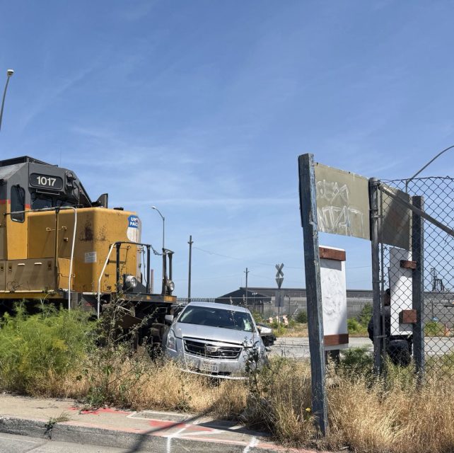 A yellow locomotive is stopped on tracks next to a silver SUV at a railroad crossing, with overgrown weeds and industrial buildings in the background.