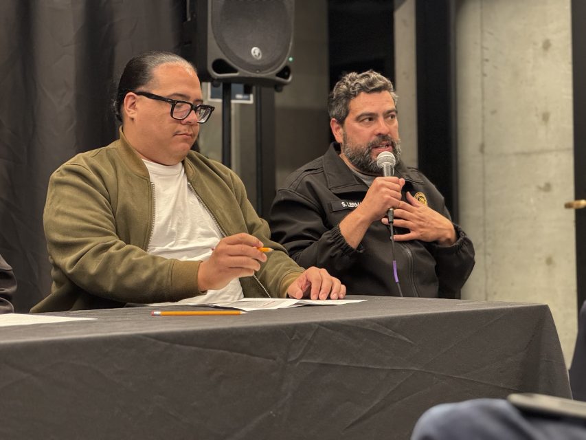 Two men sit at a table during a panel discussion; one speaks into a microphone while the other holds a pencil and looks at papers.