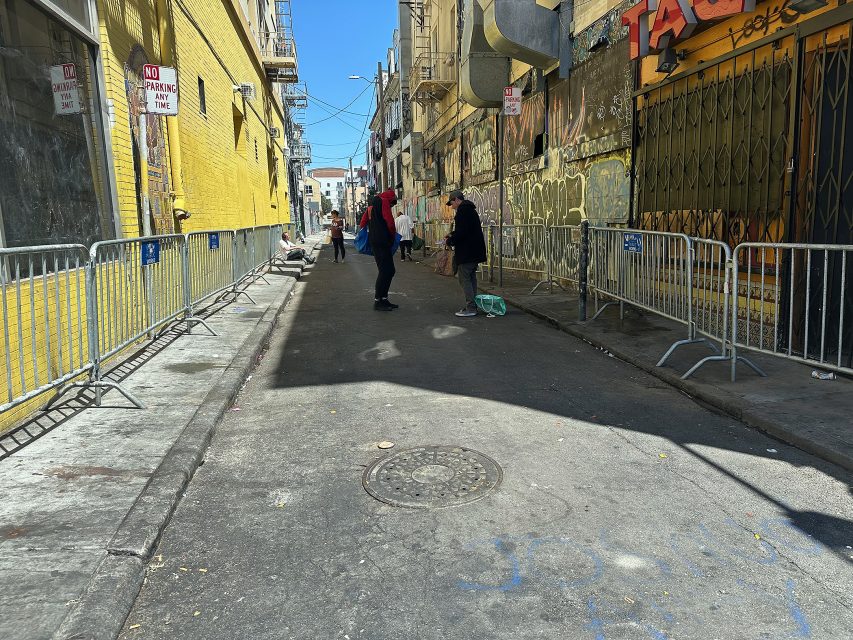 Two people stand and talk in a narrow alley lined with yellow and gray buildings, metal barricades, and graffiti, under bright sunlight.