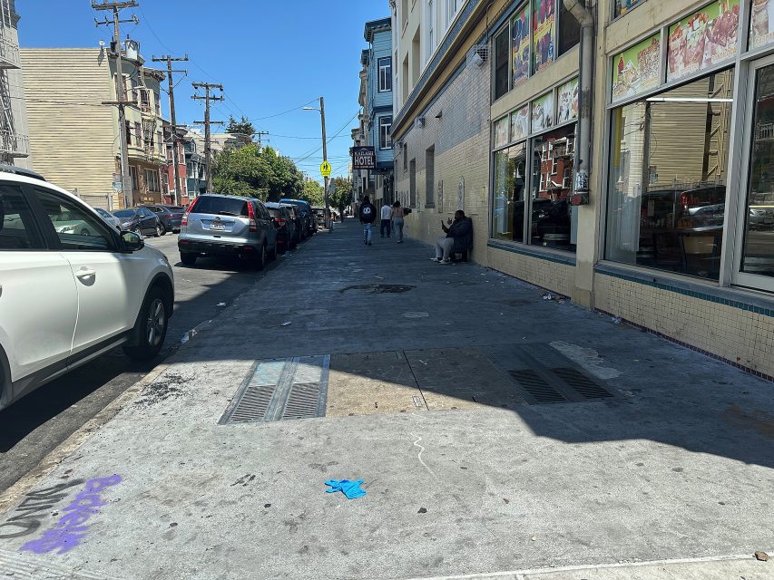 A city sidewalk with parked cars, pedestrians, and a storefront in daylight; a blue glove and some litter are visible on the ground.