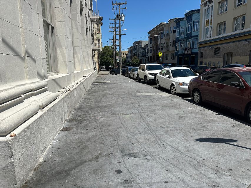 A city sidewalk runs alongside a row of parked cars and multi-story buildings on a sunny day, with shadows cast on the ground.